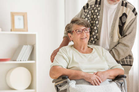 Smiling elder woman in a wheelchair looking away. Supported by her husband