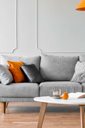 Grey sofa with pillows and a coffee table with an orange in a living room interior. Real photo
