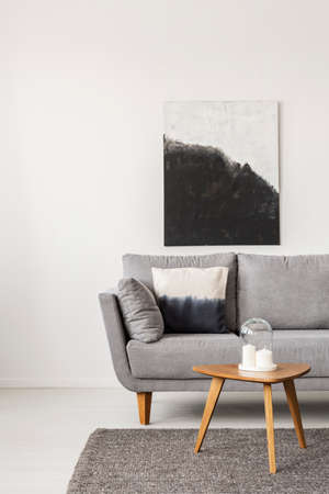 Abstract black and white painting on empty white wall with wooden coffee table with candles and grey...