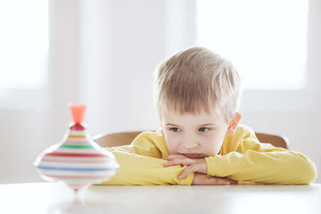 Thoughtful little boy sits at the table and looks at the spinning top