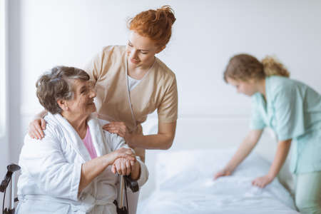 Elderly woman on wheelchair in nursing home with helpful doctor at her side and young nurse making the bedの写真素材