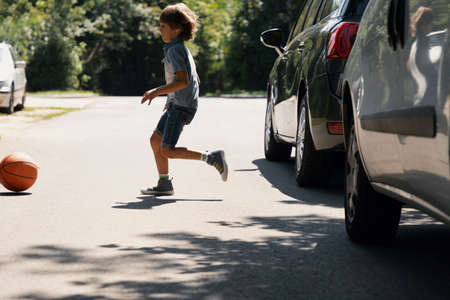 Careless boy running behind the ball on the road next to carsの写真素材