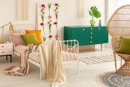 Striped blanket on comfortable single bed in classy bedroom with green wooden cabinet and wicker peacock chairの写真素材