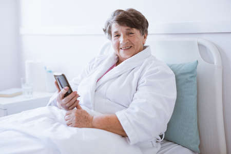 Smiling senior lady holding mobile phone while sitting in hospital bed