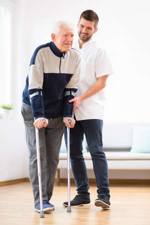 Elderly man walking on crutches and a helpful male nurse supporting himの写真素材
