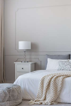 Simple white lamp on wooden night stand table with two drawers next to comfortable king size bed with cozy beddingの写真素材