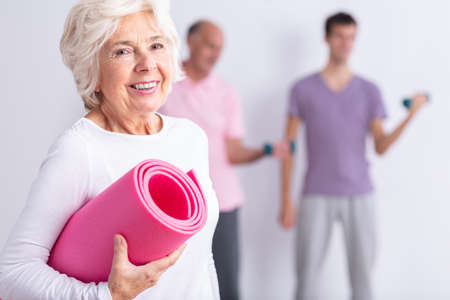 Happy senior woman holding pink joga mat, men exercising in backgroundの写真素材