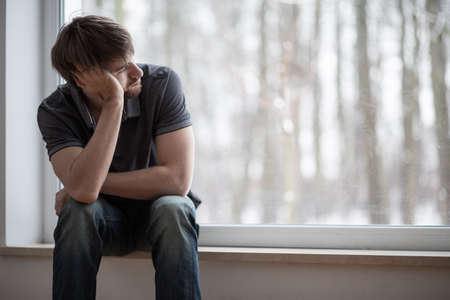 Thoughtful young man sitting on windowsill looking through the window