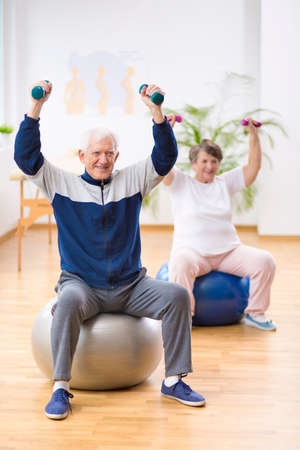 Elderly man and woman exercising on gymnastic balls during physiotherapy session at hospitalの写真素材
