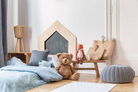 Stylish kid's bedroom in tenement house. Grey pouf on the parquet, wooden table with bags and toys, bed with blue bedding.の写真素材