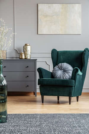 Emerald green armchair with pillow next to grey wooden commode in dark living room interiorの写真素材