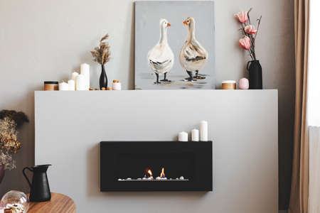 Flowers in vases, candles and rustic painting on shelf above eco fireplace in fashionable living...