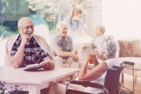 Elderly woman in the wheelchair drinking tea while talking with smiling senior manの写真素材