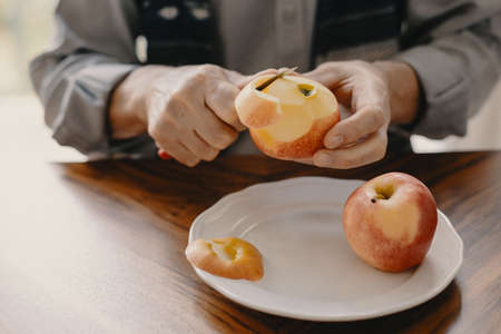 Senior caucasian man peeling an apple with a knife sitting at the wooden table