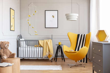 Black and white blanket on yellow egg chair in grey baby bedroom with black wooden stool with mug and grey crib with cozy blanketの写真素材
