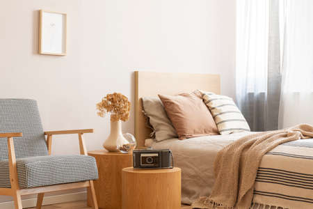 Vintage camera on round wooden table in the middle of elegant bedroom with retro armchair and single bed with beige bedding and flowers in vaseの写真素材