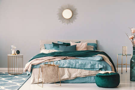 Clock and flower in fancy vase on nightstand table next to king size bedroom with emerald green and beige beddingの写真素材