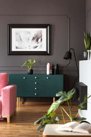 Poster in a black frame above a dark turquoise dresser and plants in a gray living room interior...