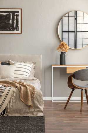 Elegant round mirror in wooden frame above fancy console table with flowers in vase in trendy bedroom interior with beige vaseの写真素材