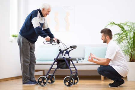 Elderly grandfather with walker trying to walk again and helpful male nurse supporting himの写真素材