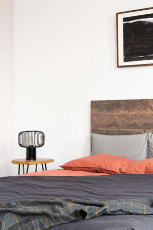 Closeup of lamp on wooden nightstand and black poster on the white wall of elegant bedroom interior with king size bed with wooden headboardの写真素材
