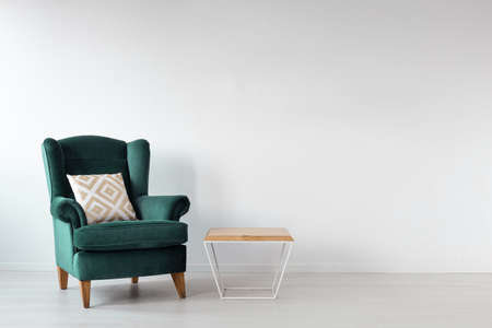 Green armchair with cushion next to wooden table in white loft interior with copy space. Real photo