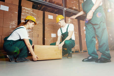 Two young warehouse workers in dark green uniforms and yellow helmets lifting heavy packageの写真素材