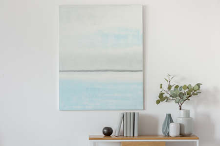 Pastel blue and white abstract oil painting on empty white wall with console table with flowers in...