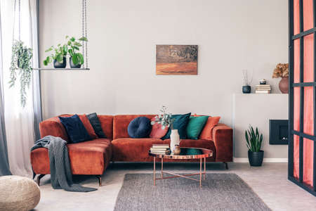 Real photo of colorful pillows on a red corner couch in white living room interior with gray rugの写真素材