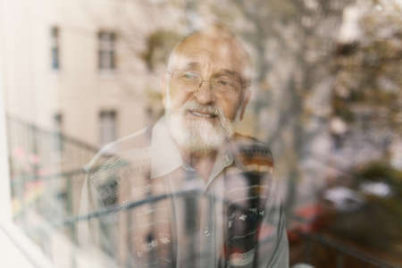Smiling senior man looking through the window and waiting for his familyの写真素材