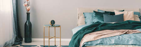 Flowers in bottle shape vase next to clock on stylish nightstand with golden legs next to king size...