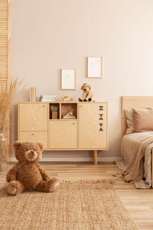 Cute brown teddy bear sitting on the floor of beige bedroom interior with wooden commodeの写真素材
