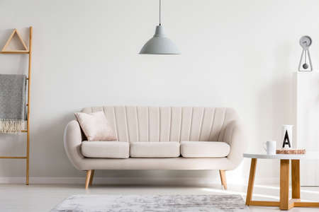 Grey lamp above pillow on white sofa next to wooden coffee table with mugs, copy space on empty wallの写真素材