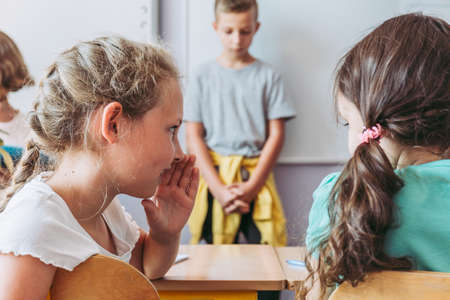 Two young girls gossiping during classes about the sad boy standing by the boardの写真素材