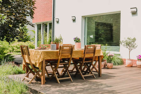 Dining table covered with orange tablecloth standing on wooden terrace in green gardenの写真素材