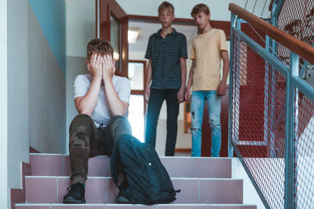 Sad kid with backpack sitting on stairs at school, two classmates behind himの写真素材