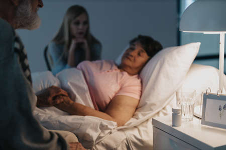 Senior man sitting by his sick wife's bed and holding her handの写真素材
