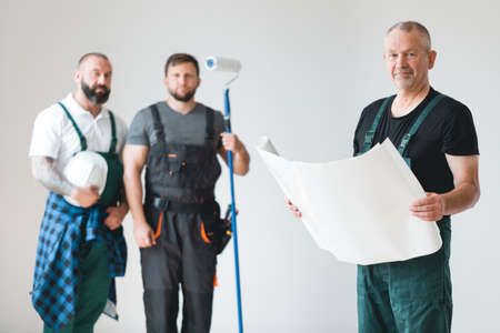 Crew of three professional builder with painting roll and renovation plans standing in empty interiorの写真素材