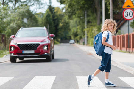 Young boy with a backpack goes through the pedestrian crossing, red cars let him throughの写真素材