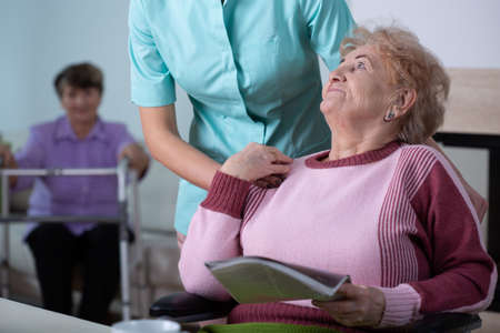 Senior lady reading a newspaper in common room of professional nursing home with helpful social worker supporting herの写真素材