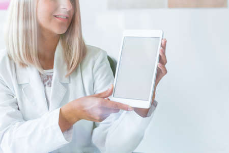Pretty blonde nurse in medical uniform with tablet, copy space on empty white wall