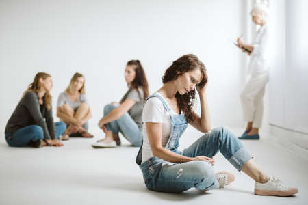 Worried woman sitting alone on the floor in a white room during group psychotherapy