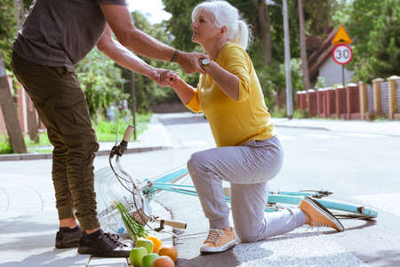 Handsome man helps an elderly woman get up after falling off a bicycle on a suburban roadの写真素材