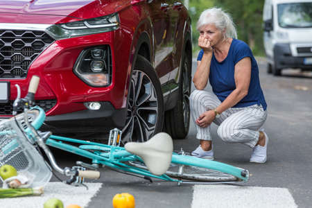 Worried old lady kneels beside her red car after she hits a bikerの写真素材