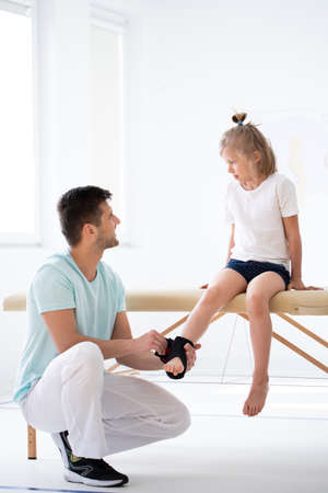 Cute little injure girl on massage table exercising with young male doctorの写真素材