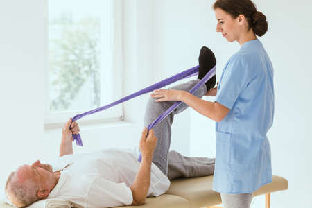 Injured patient lying on a physiotherapy bed and performing exercises with tapes under the care of a doctorの写真素材