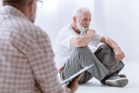 Sad gray senior man sitting on the floor during group psychotherapy for menの写真素材