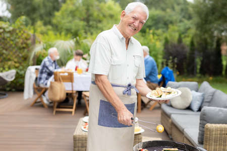 Smiling senior man wearing apron and holding plate of food during garden hooplaの写真素材