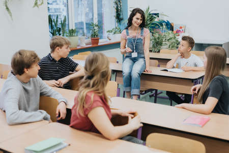 Young teacher in denim overalls sits on a bench and advises after-school workshops at schoolの写真素材