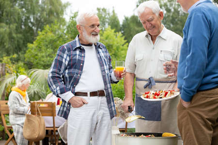 Senior friends with drinks and food standing next to grill during garden partyの写真素材
